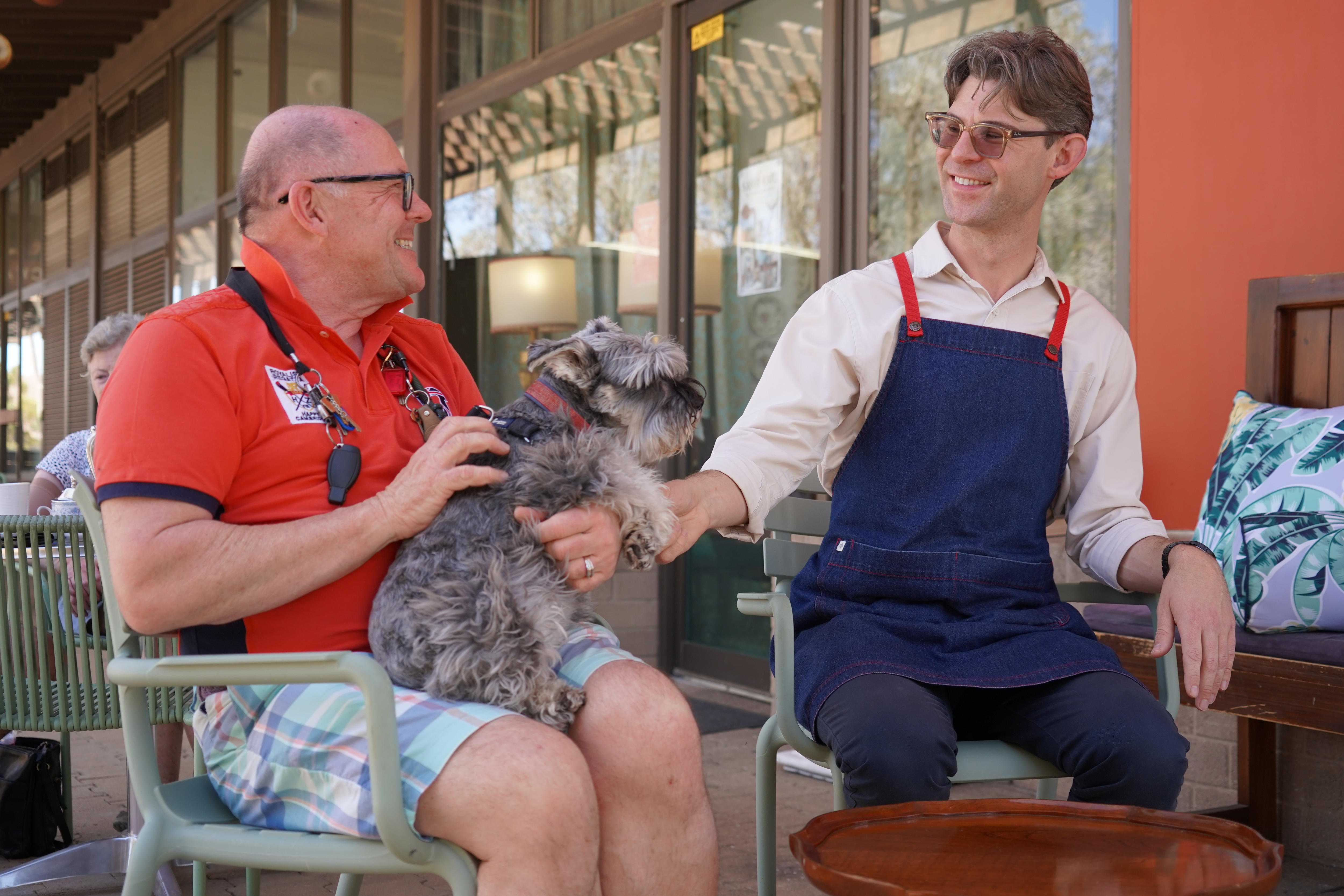 Two men sit outside a cafe smiling at each other. One is holding a dog on his lap, the other wears an apron. 