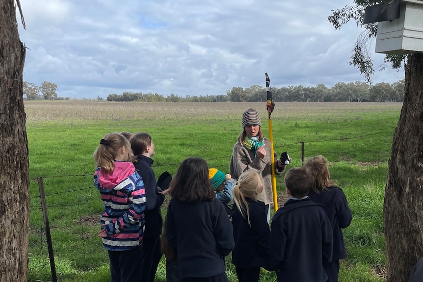 An adult speaks to a group of children standing near some trees.