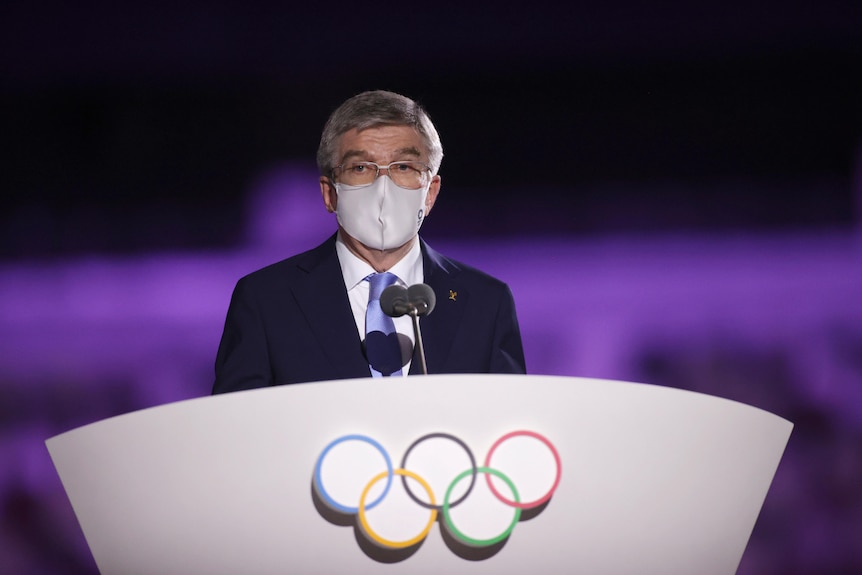 IOC president Thomas Back stands behind a lectern with the olympic rings on it, wearing a suit with a blue tie and a grey mask