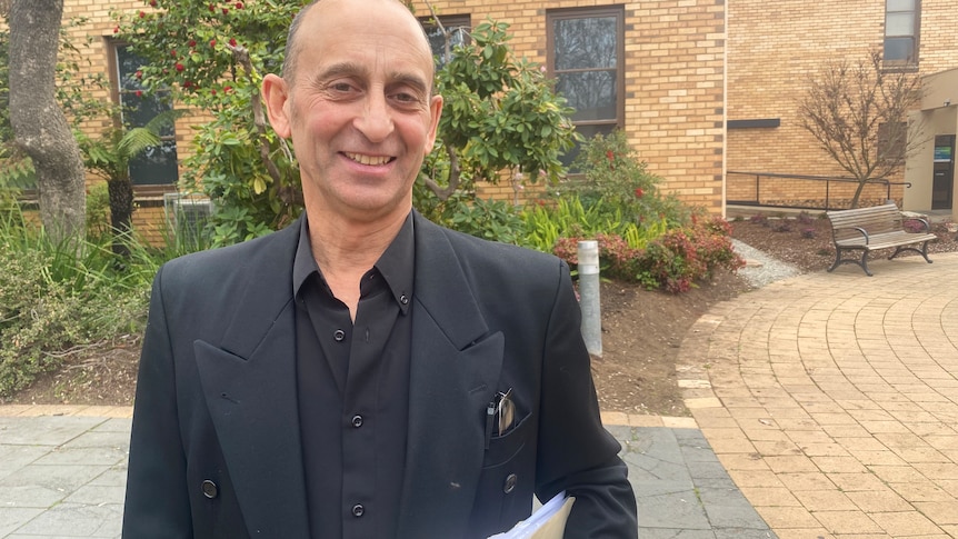 A bald man in dark formal wear, holding documents and smiling outside a courthouse.