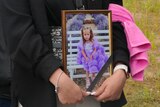 A woman holds a portrait of a little girl in a purple dress. 