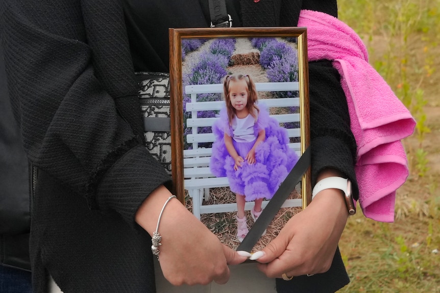A woman's hands clutch a photo frame with a picture of 4-year-old Liza, dressed in purple, who was killed in Ukraine. 