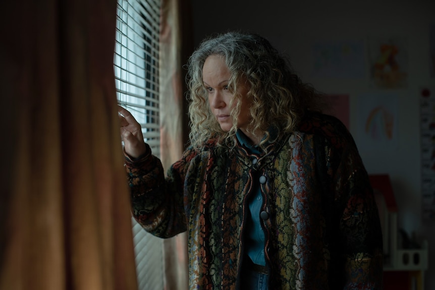 A woman with curly grey hair looks through the blinds of a window.