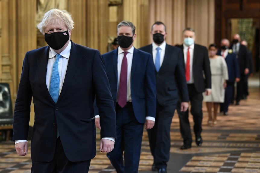 A line of men and women in suits and masks walks past the camera.