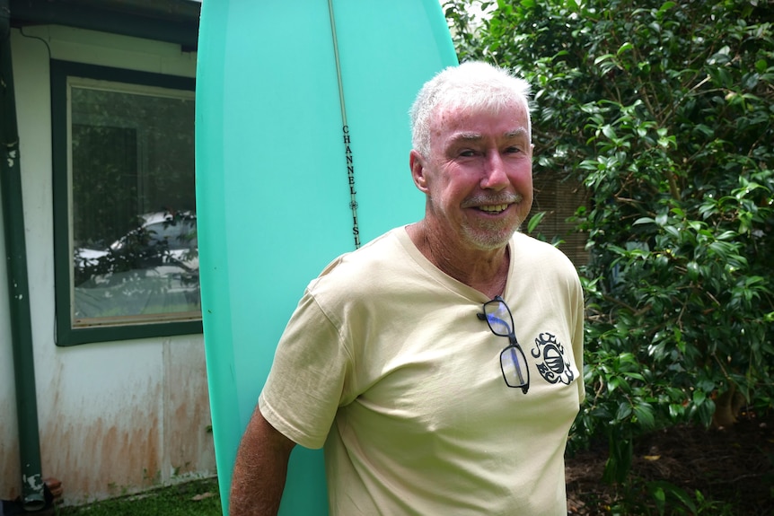 White-haired man wearing yellow shirt smiling in front of aqua blue surfboard.