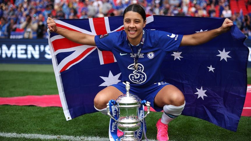 Sam Kerr squats behind the Women's FA Cup while holding the Australian flag and wearing her Chelsea jersey.