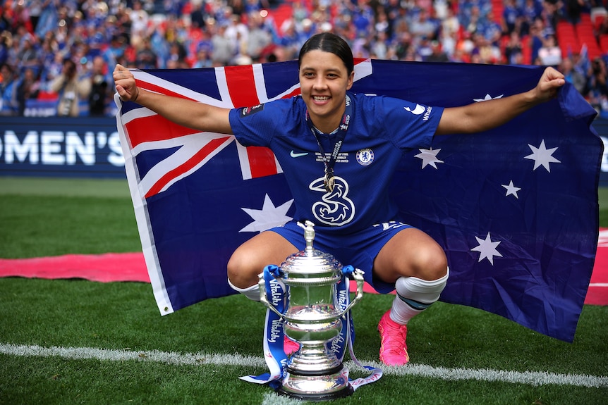 Sam Kerr squats behind the Women's FA Cup while holding the Australian flag and wearing her Chelsea jersey.