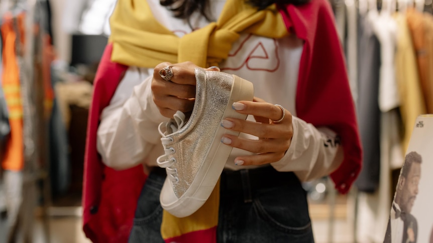 Woman holding a white, glittery sneaker in front of the camera, with a red and yellow sweater tied over her shoulders.