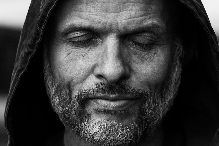 Black and white portrait of a man with a grey beard wearing a hoodie. His eyes are closed and he has a slight smile.