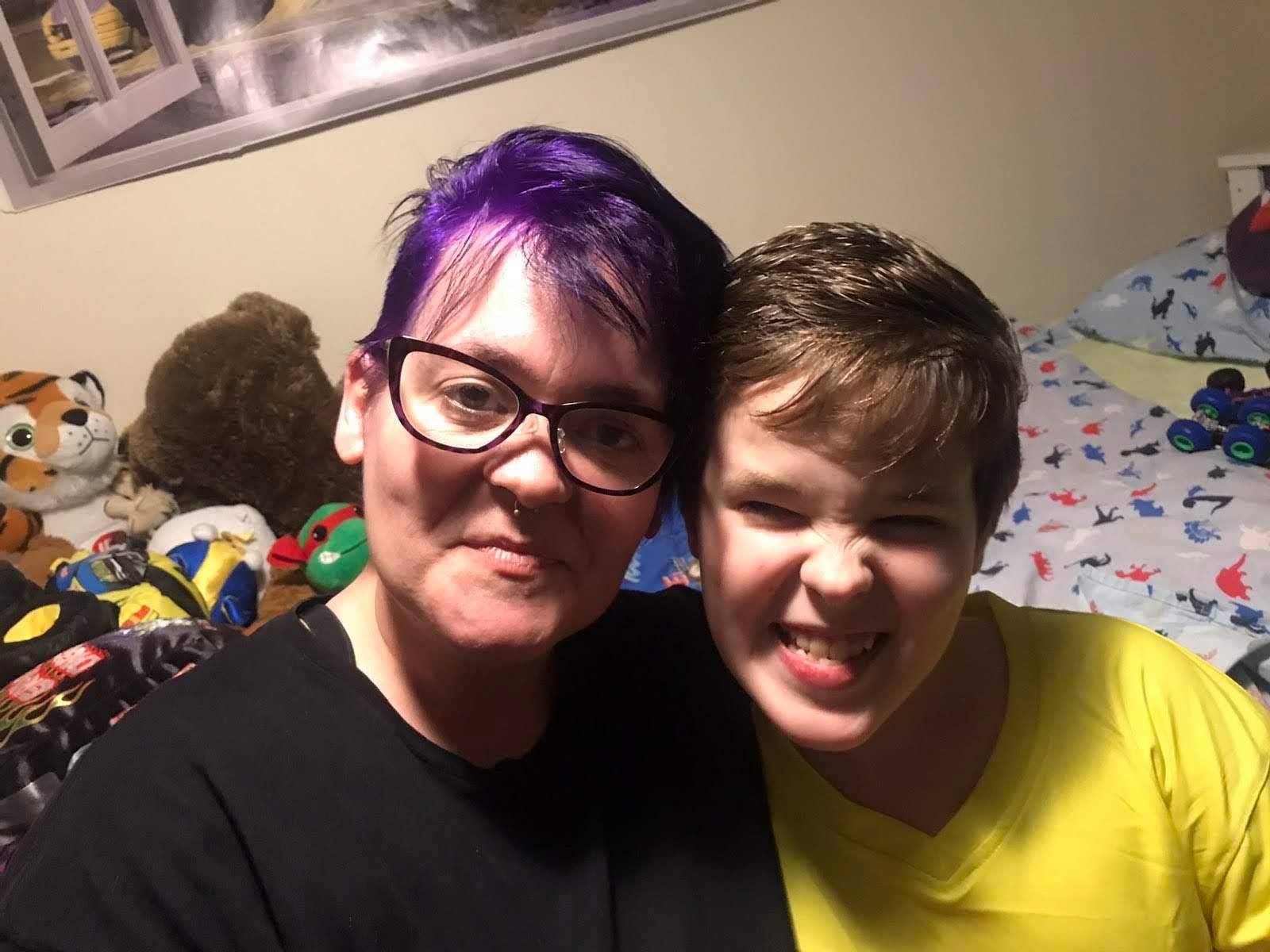 A woman with short purple hair smiles beside a teen boy pulling a funny face.