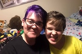 A woman with short purple hair smiles beside a teen boy pulling a funny face.