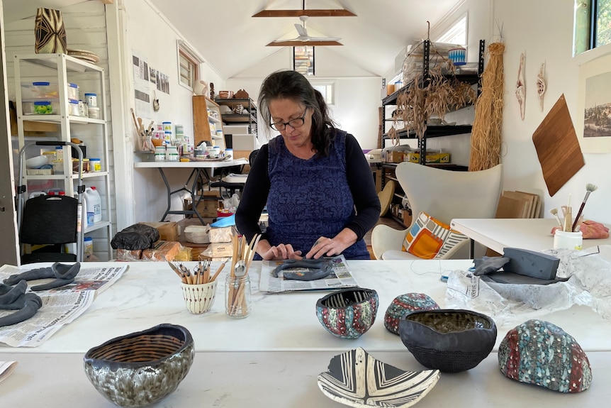 A 50-something woman with grey hair and glasses is at work in her studio, standing at a table making sculptural and pottery work