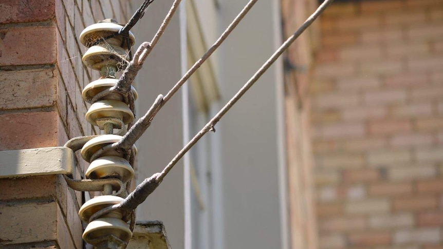 An set of electricity wires are attached to a cobwebbed metal rod mounted on the side of a brick building.