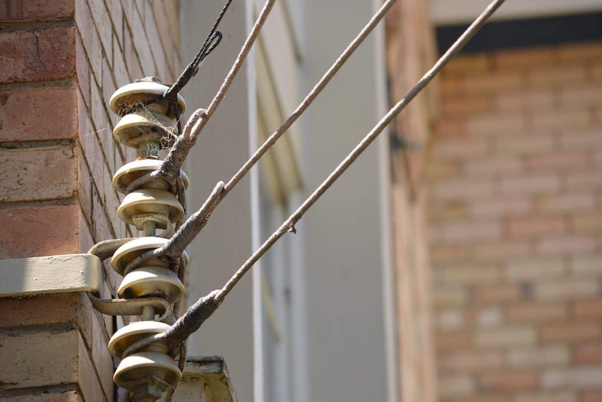 An set of electricity wires are attached to a cobwebbed metal rod mounted on the side of a brick building.