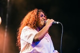 A woman with long dark hair and wearing a white top smiles while holding a microphone on stage
