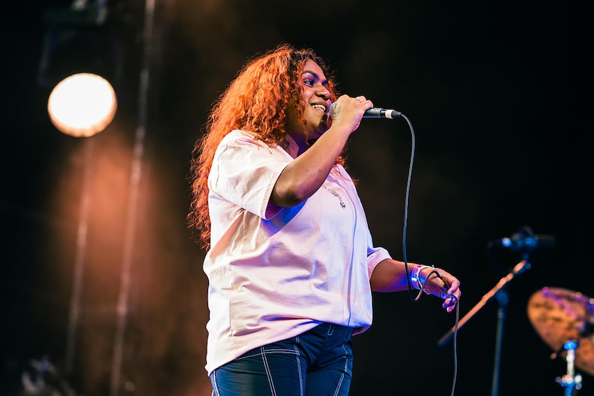 A woman with long dark hair and wearing a white top smiles while holding a microphone on stage