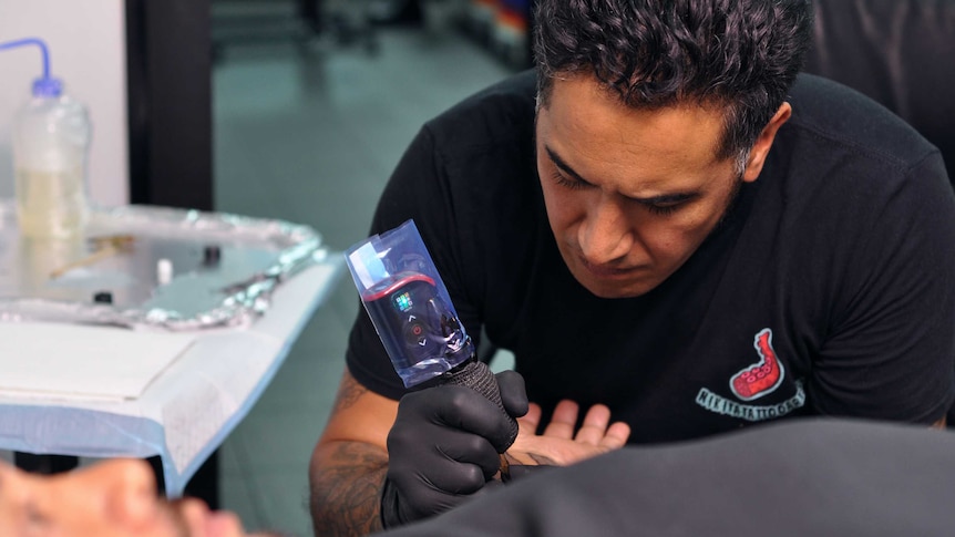 A Maori man tattoos a traditional Moko tattoo onto the arm of a client.
