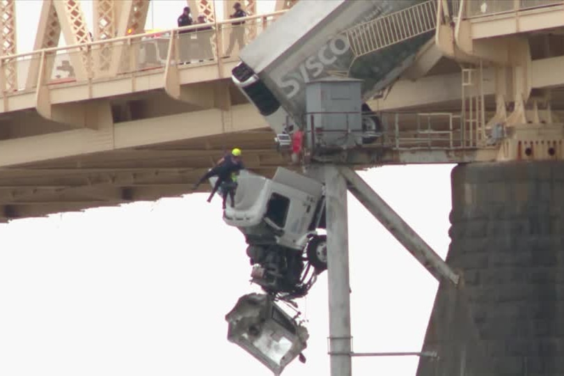 A zoomed in photo of a semi-trailer truck and a rescue worker on a line dangling over the side of a large bridge.