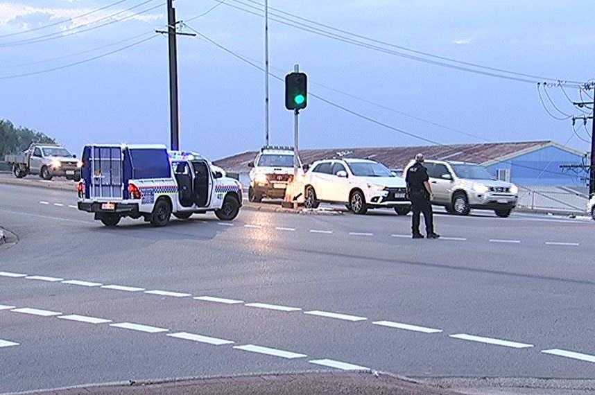 Two police vehicles block a two-way road with a police officer standing in the middle of the street. The sky is light above.