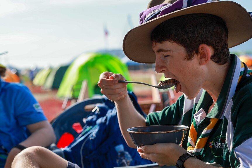 A young lad in a scout uniform enjoys a bowl of baked beans at a campsite.