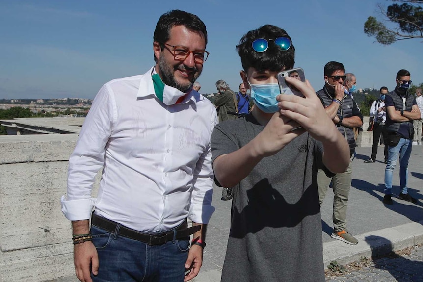 Matteo Salvini, with a face mask in the Italian flag colours worn under his chin, stands next to a teen taking a selfie