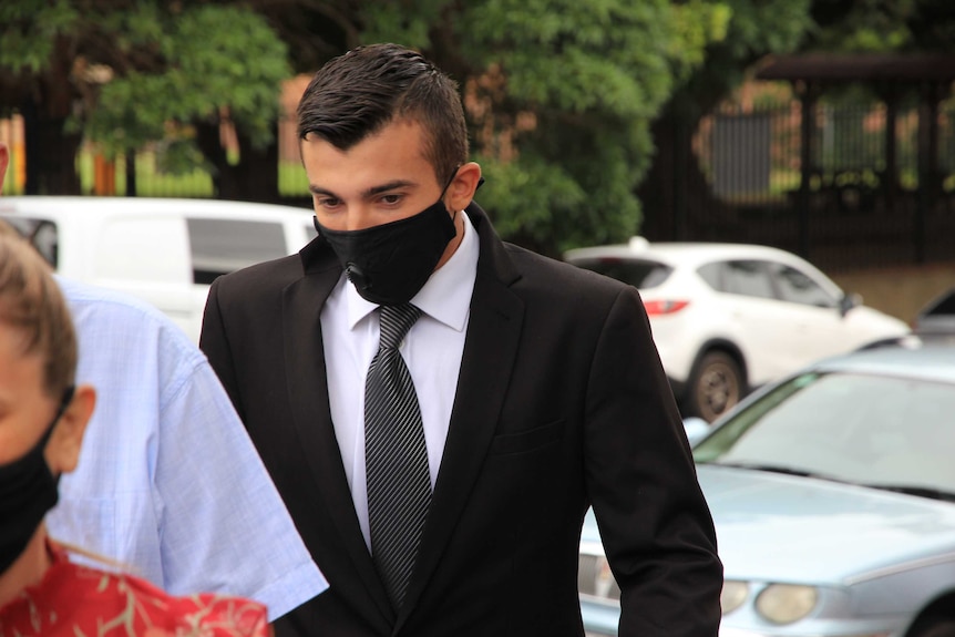 A man wearing a black suit and face mask walks along a city street.