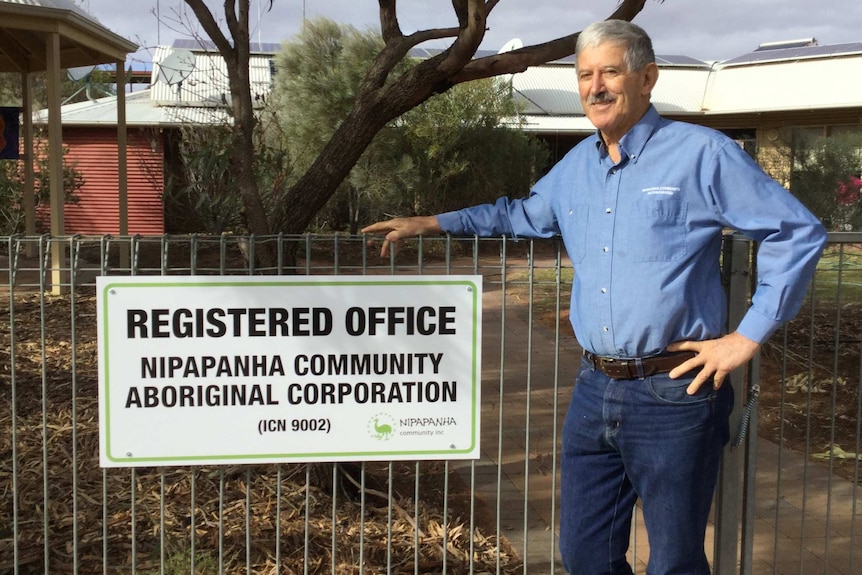 A man with grey hair wears a blue shirt in front of a building.