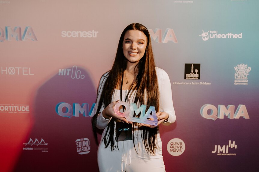An image of Cloe Terare in white at QMAs holding award in front of media wall with QMA logo