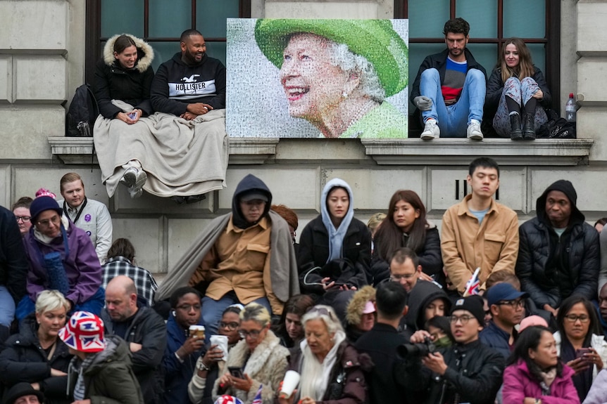 Crowds sit on the ground and on windowsills, with a portrait of Queen Elizabeth in the centre. 