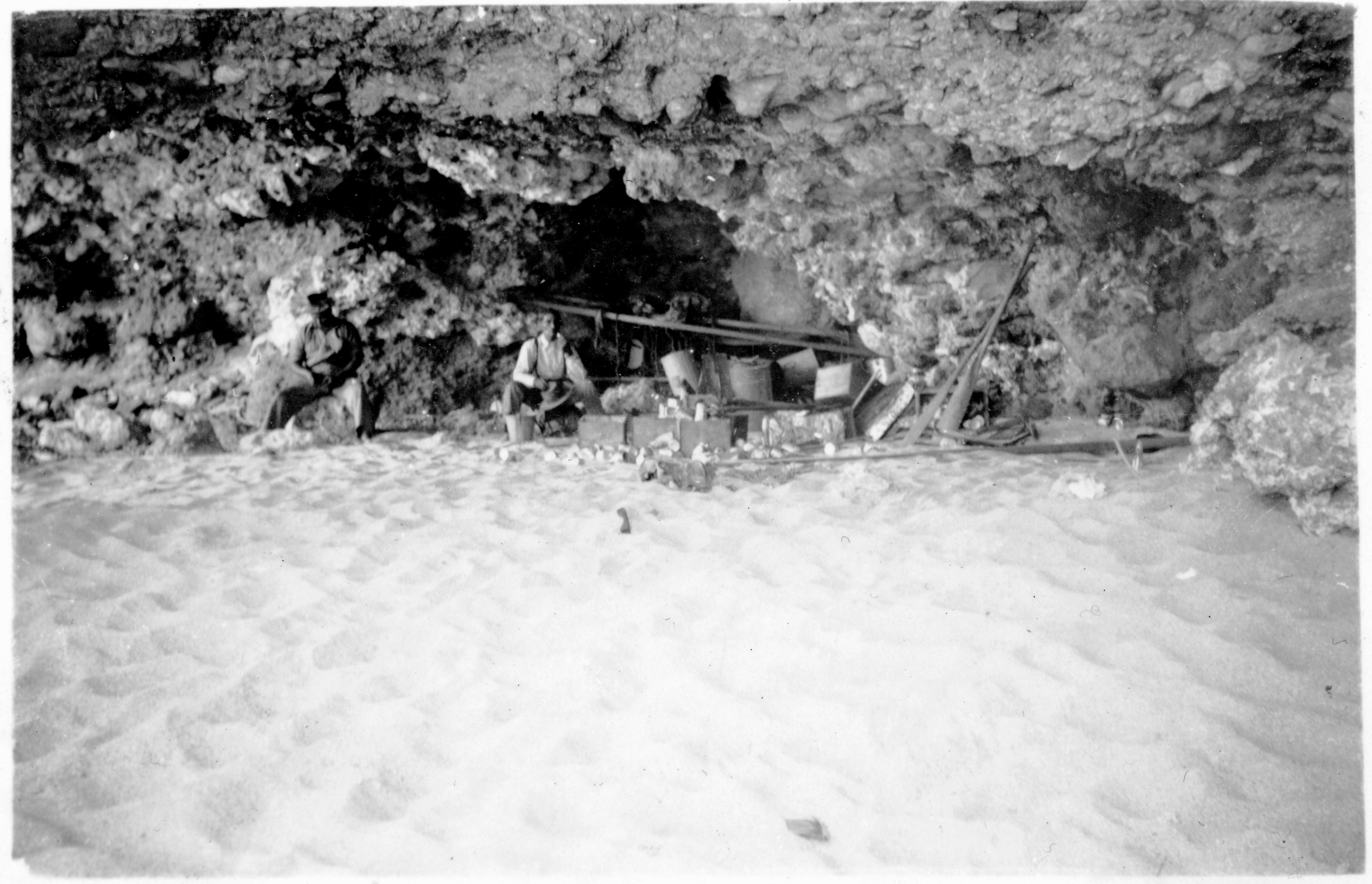 An old photo of a man surrounded by equipment stores in a low cave.