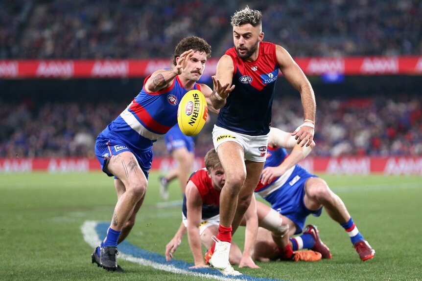 A Melbourne Demons AFL player reaches for the ball as a Western Bulldogs opponent tries to tackle him.