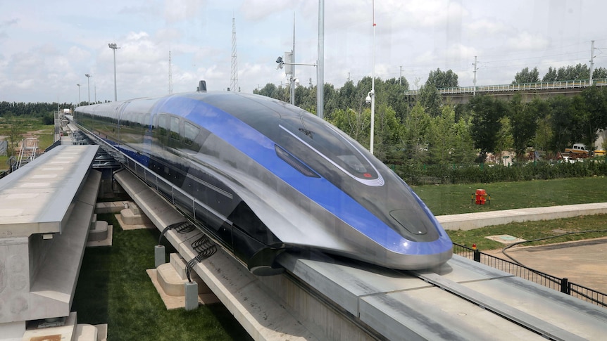 The long, slanted nose of a blue, grey and black bullet train sits on a special concrete track wit one metal rail.