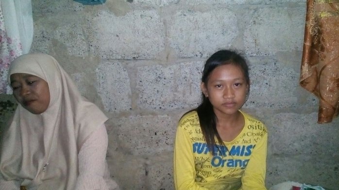 Reski Suci Ramdani, wearing a yellow t-shirt, sits next to someone in front a wall.