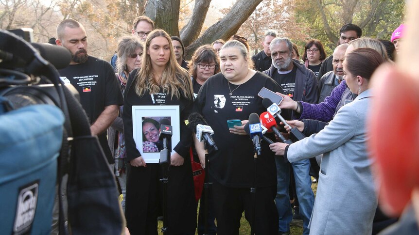Apryl Watson, holding a photo of her mother, stands next to her sister Belinda Stevens as they speak to media.