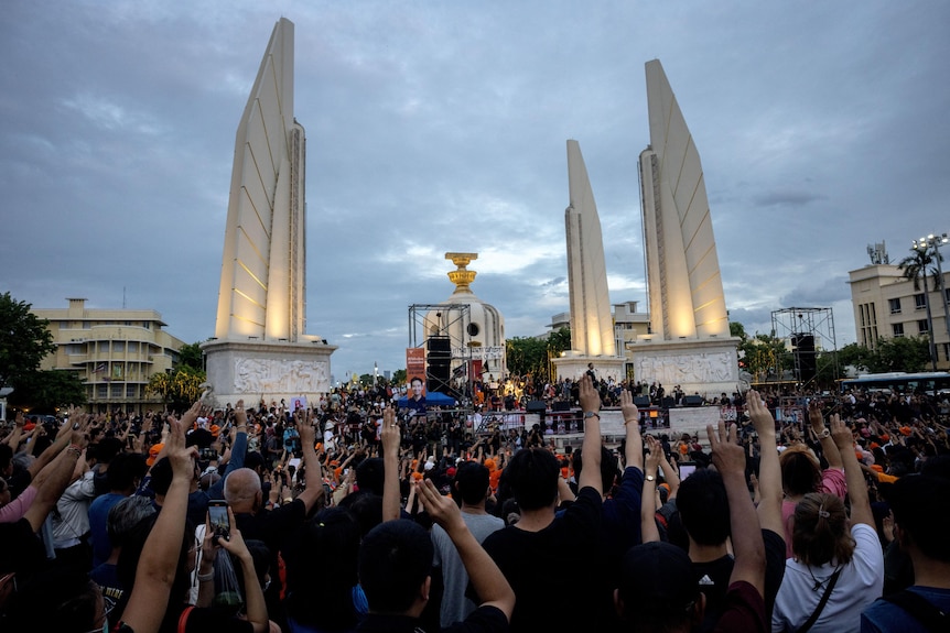 A large group of people hold three fingers in the air, surrounding a monument
