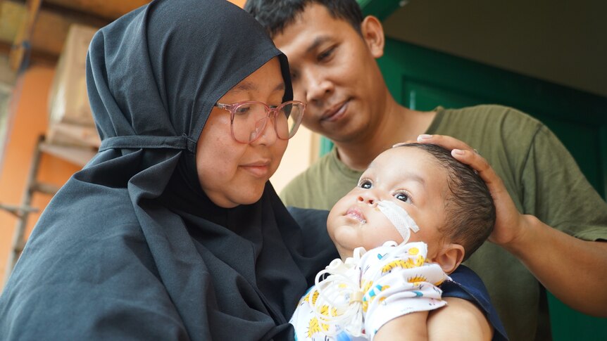 Indonesian woman holding her infant son with her husband standing next to her.