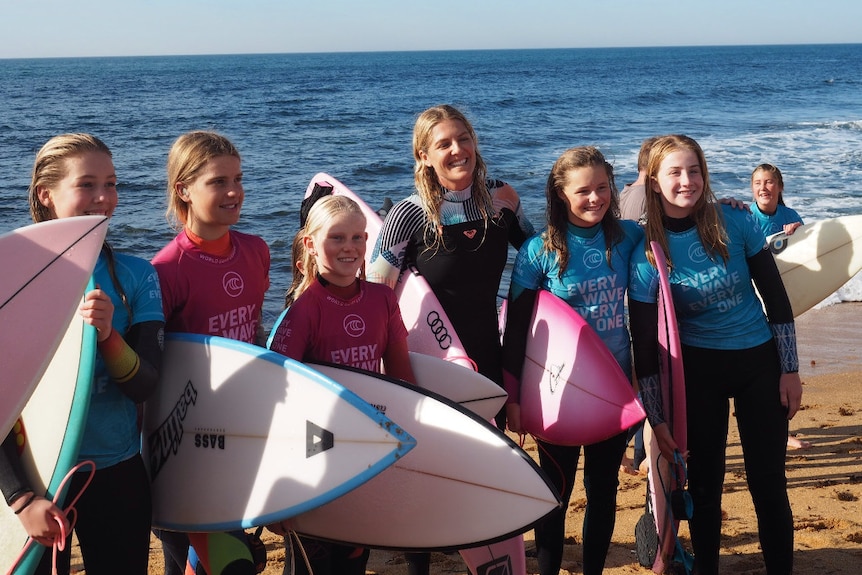 A professional surfer poses for a photo with five junior female surfers on a beach wearing wetsuits and carrying boards.