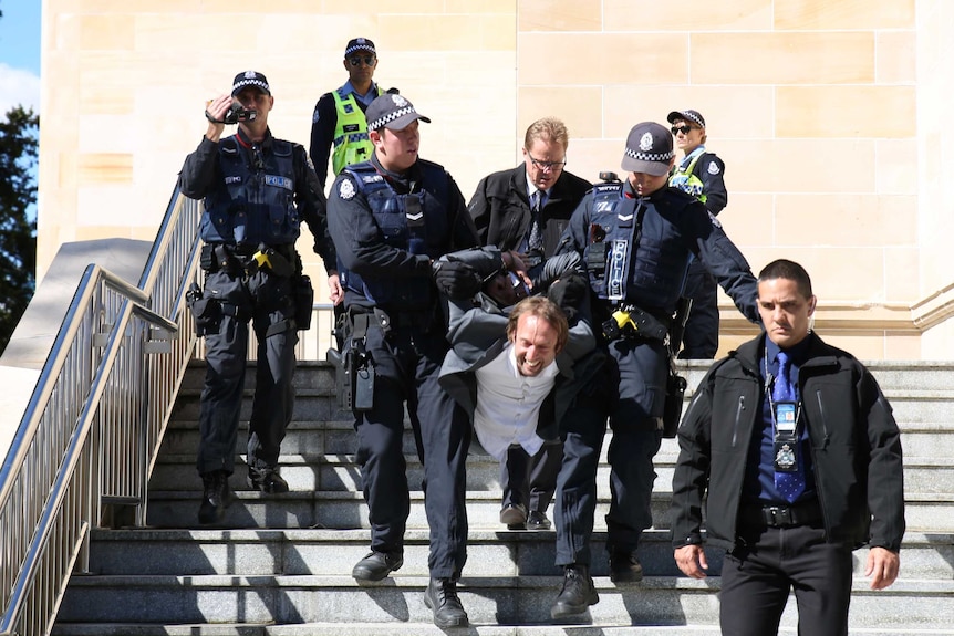 A man is carried down steps with his hands behind his back by armed police officers.