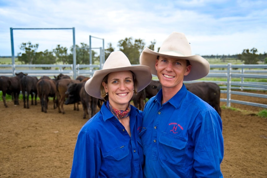 A smiling couple in big hats pose in front of beef cattle in a cattle yard.