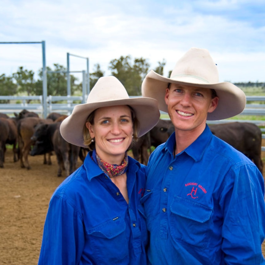 A smiling couple in big hats pose in front of beef cattle in a cattle yard.