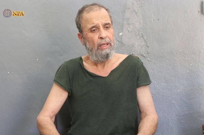 A man in a green T-shirt is seen sitting against a grey concrete wall.