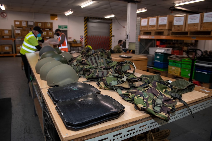 Военнослужащие в светоотражающей одежде упаковывают ящики на складе.  Шлемы и камуфляжные бронежилеты раскладываются на столе.