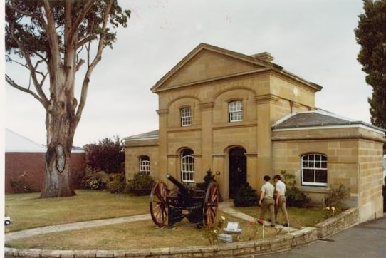 Photo of sandstone building with canon at front. Tree in left corner with concrete in it.