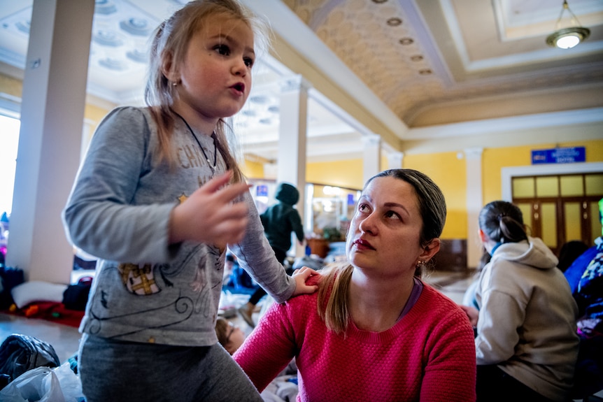 A young girl stands and talks inside a train station, with her left hand placed on her sitting mother's right shoulder.