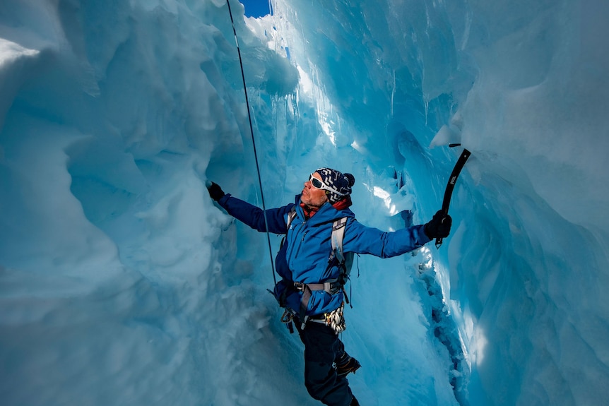 A man uses an ice axe to climb the bumpy wall of a blue crevasse.