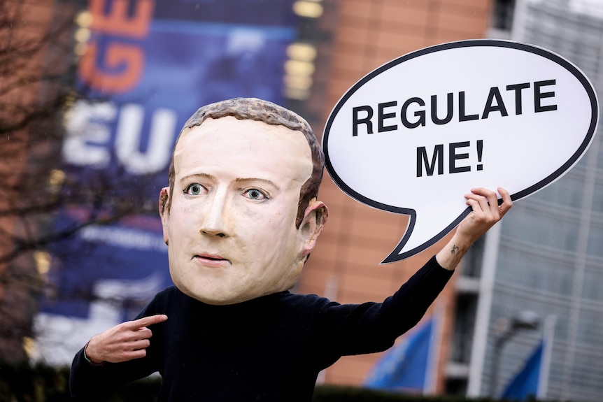 An activist wearing a mask depicting Facebook CEO Mark Zuckerberg holds a banner reading "Regulate me"