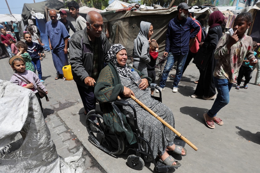 An elderly man pushes a woman in a wheelchair through a crowd of refugees.