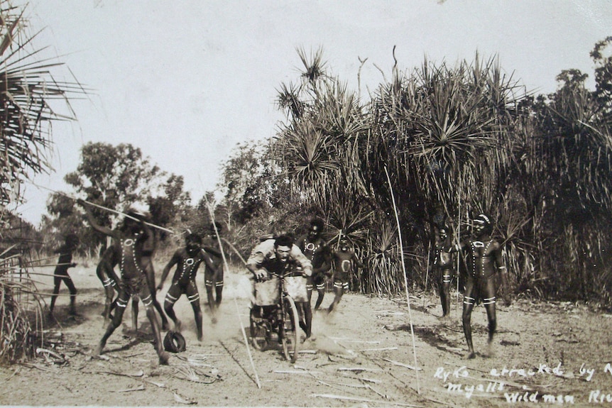 A man on a bike rides between Aboriginal men with spears.