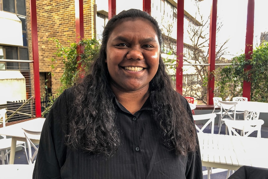 A young Indigenous girl wearing a black shirt, standing in an open cafe smiles at the camera. 