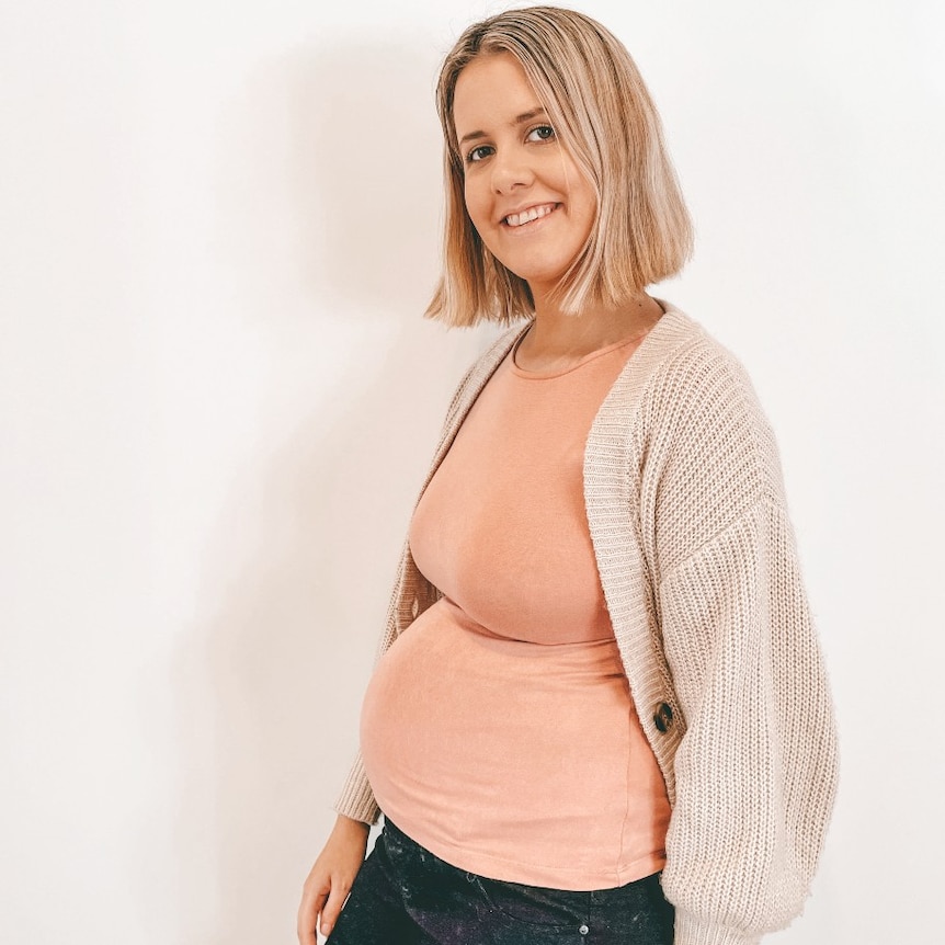 A pregnant lady with blonde hair standing in front of a white wall and smiling at the camera